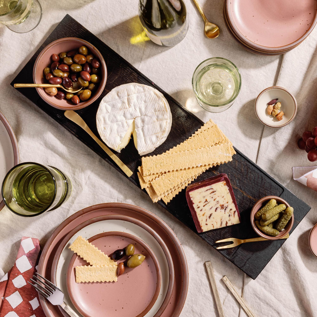 Cheese board with olives, crackers, brie, pickles, and wine glasses on a linen table, surrounded by pink plates and utensils.