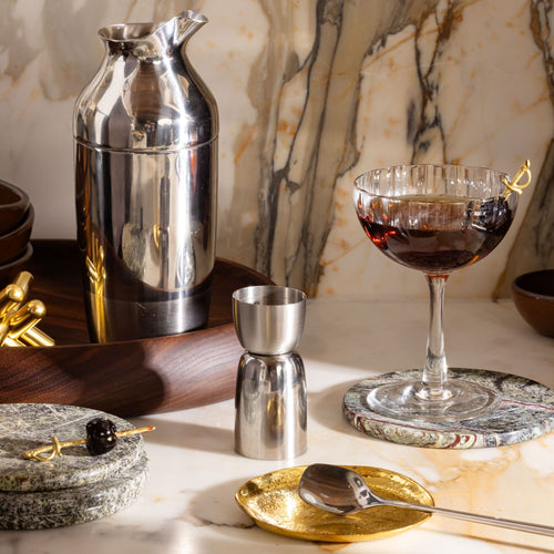 Cocktail setup with silver shaker, jigger, coupe glass with cherry garnish, and gold and stone coasters on marble.