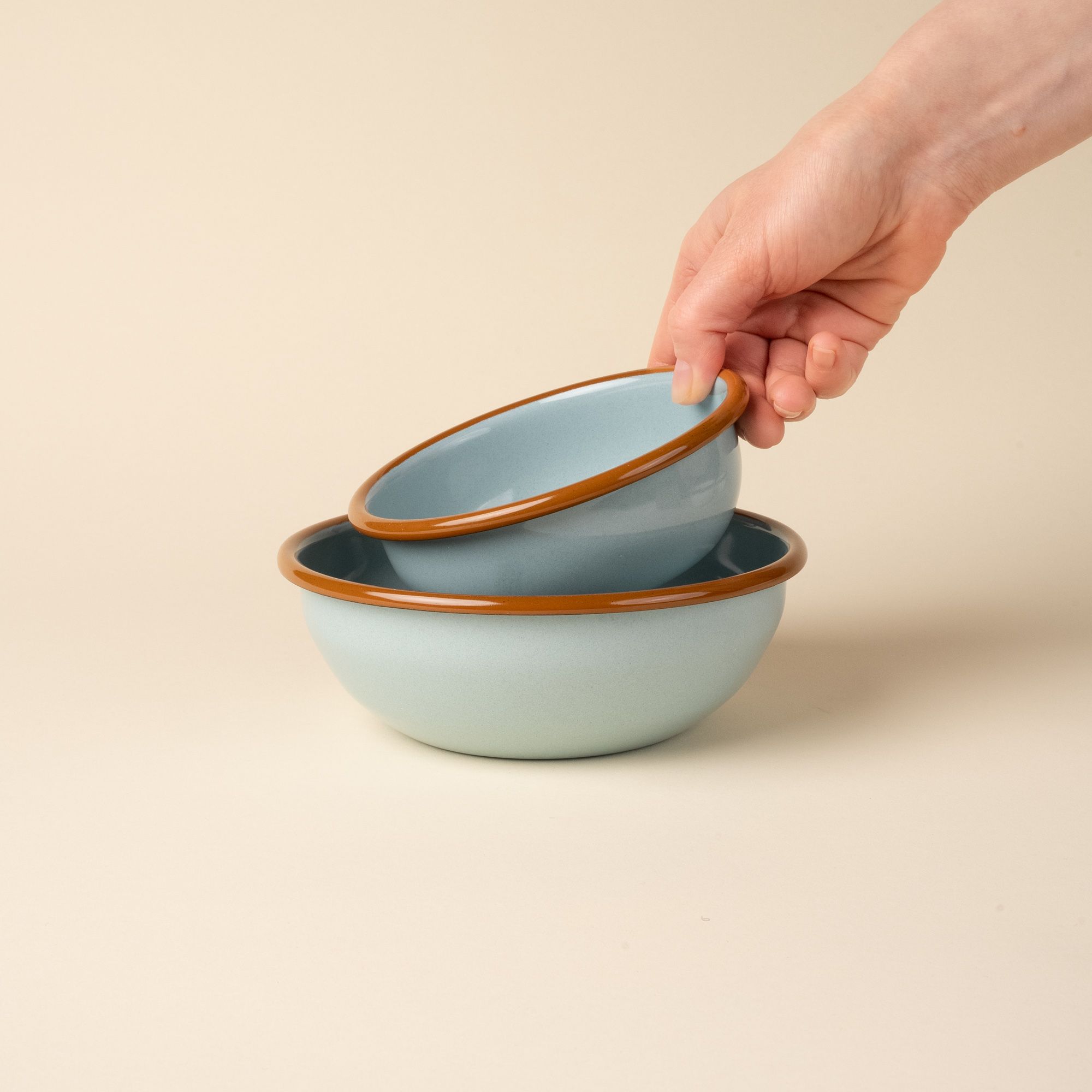 A hand holds a small enamel bowl to nest in a medium enamel bowl; both bowls are robin's egg blue with a warm brown rim.