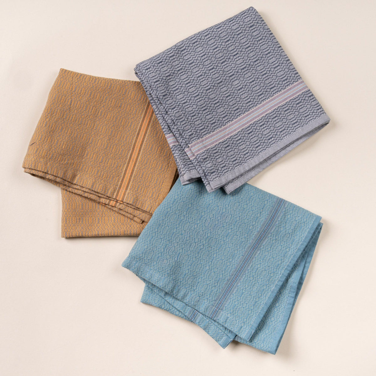 Three folded woven kitchen towels in mustard, gray, and teal, styled on a clean neutral background with subtle striped detailing.