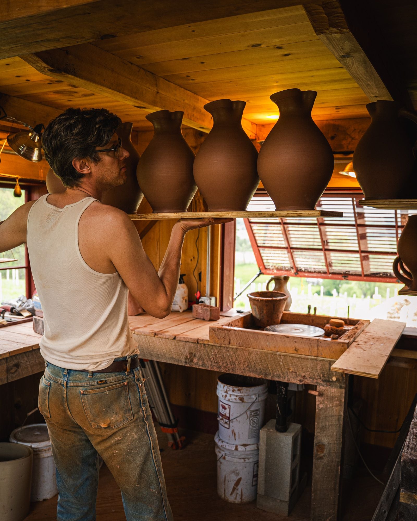 John inside his studio holding a plank of multiple wheel thrown vases