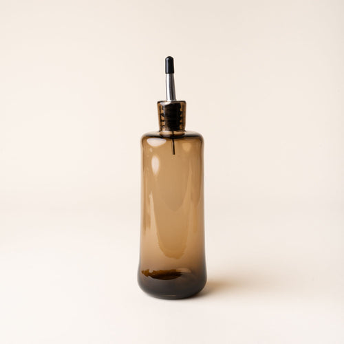 Amber glass oil dispenser with a metal spout and black cap, standing upright against a neutral background.