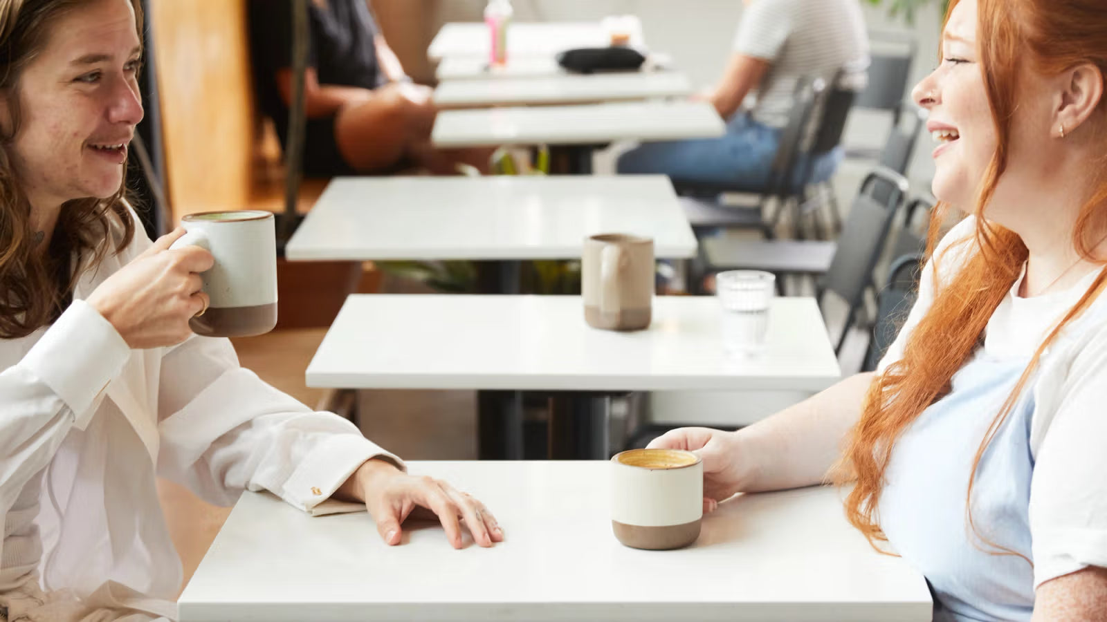 Two people seated at a café table, smiling and holding ceramic mugs with a white and brown glaze. The setting is casual, with other diners in the background.