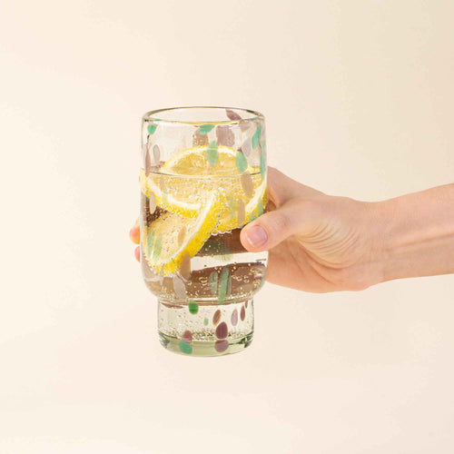 A hand holds a tall clear glass with pastel green and pink speckled patterns, filled with water and lemon slices.
