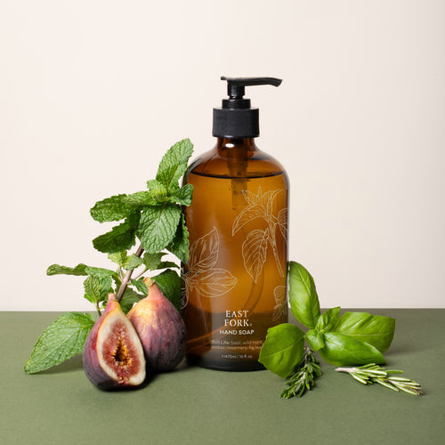 A brown glass bottle of East Fork Hand Soap labeled “Still Life” is surrounded by fresh basil, mint, rosemary, and figs on a green and cream backdrop.