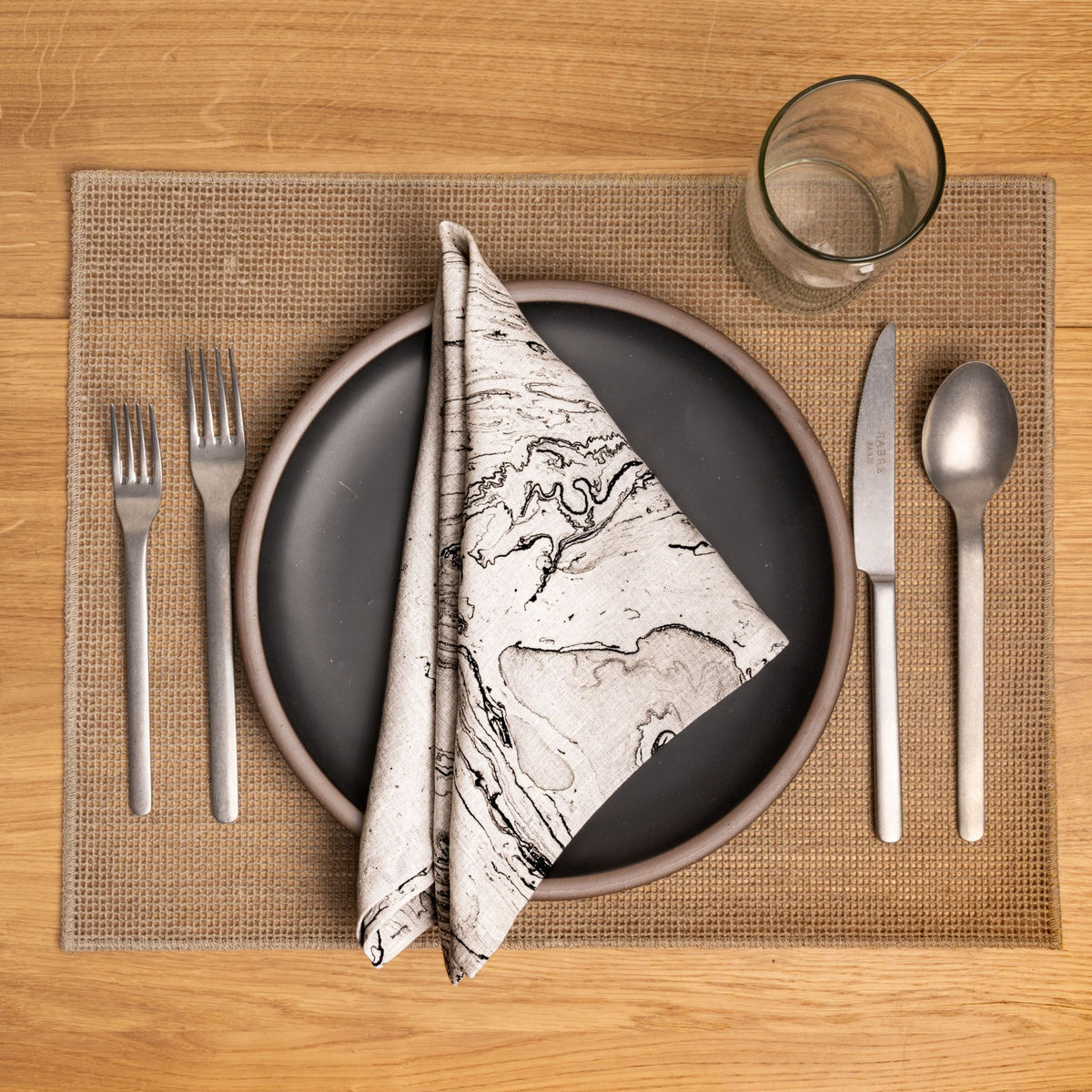 Place setting with stacked plates, gray marbled napkin, silverware, and glass on a woven tan placemat.