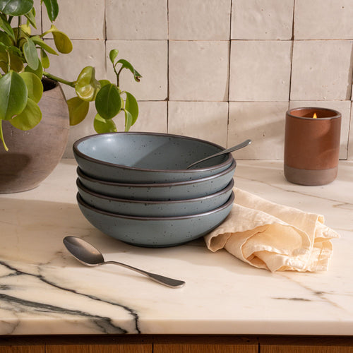 Stack of blue-grey ceramic bowls with spoons on a marble countertop beside a brown candle, beige cloth, and potted plant against tiled wall.