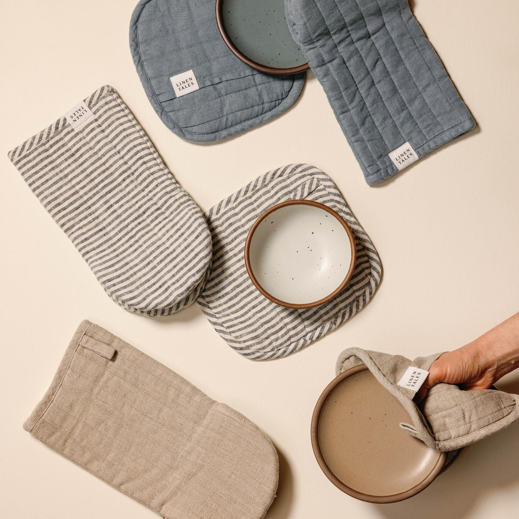 Quilted linen potholders and oven mitts in neutral beige, dusty blue, and navy-and-white stripes, each with a small ‘LINEN TALES’ label, arranged overlapping on a light background, with cake plates in matching colors.