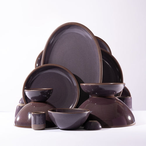 Ceramic dinnerware plates, bowls, cups, and platters of varying sizes artfully arranged in a gumdrop shape, all in a purple color with variation and glowing sheen.