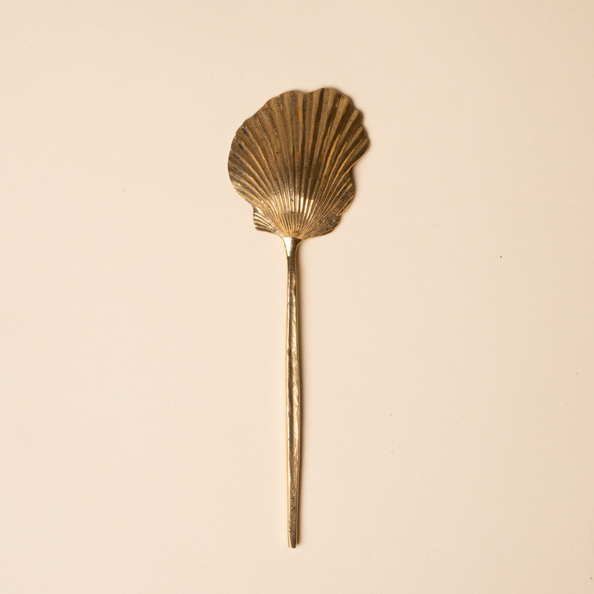 The back of a brass spoon with a scalloped shell-shaped bowl and long handle, set against a neutral beige background.