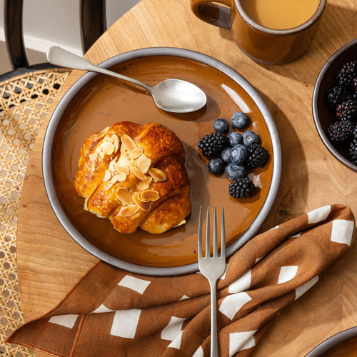 Glossy caramel brown plate with almond croissant, fresh blackberries, and blueberries, next to a folded brown gingham napkin and silverware.