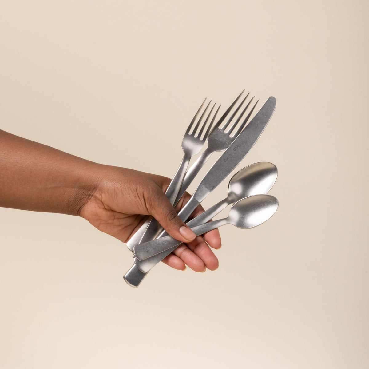 Hand holds out five matte stainless steel utensils - salad fork, dinner fork, knife, tablespoon, and teaspoon.