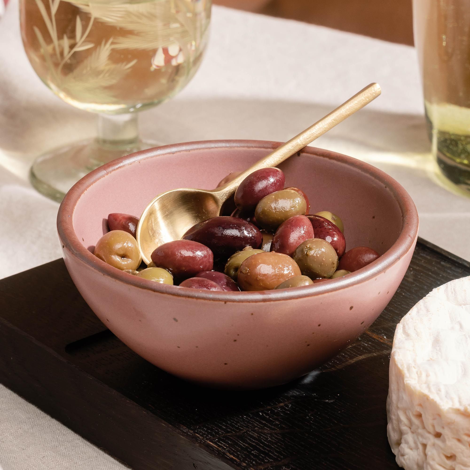 Small dusty mauve pink ceramic bowl filled with olives and a brass spoon, set on a wooden serving board with wine glasses nearby.