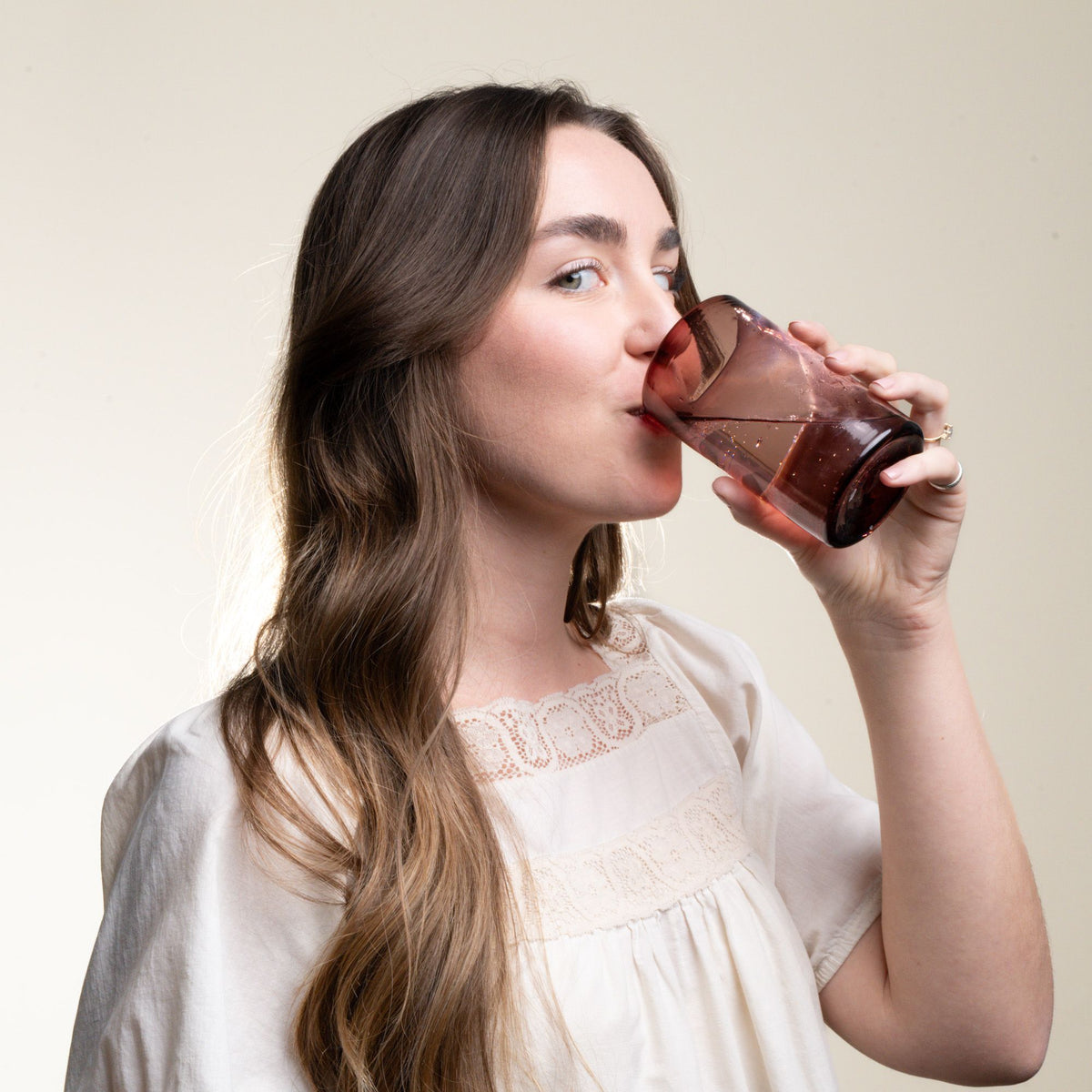 A woman staring at the camera and drinking water from a tall soft purple tumbler glass.