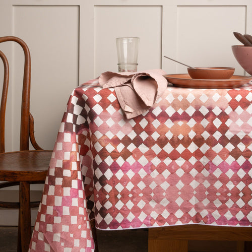Table set with pink ceramic dishware on a watercolor diamond patterned tablecloth, with a wooden chair and linen napkin nearby.