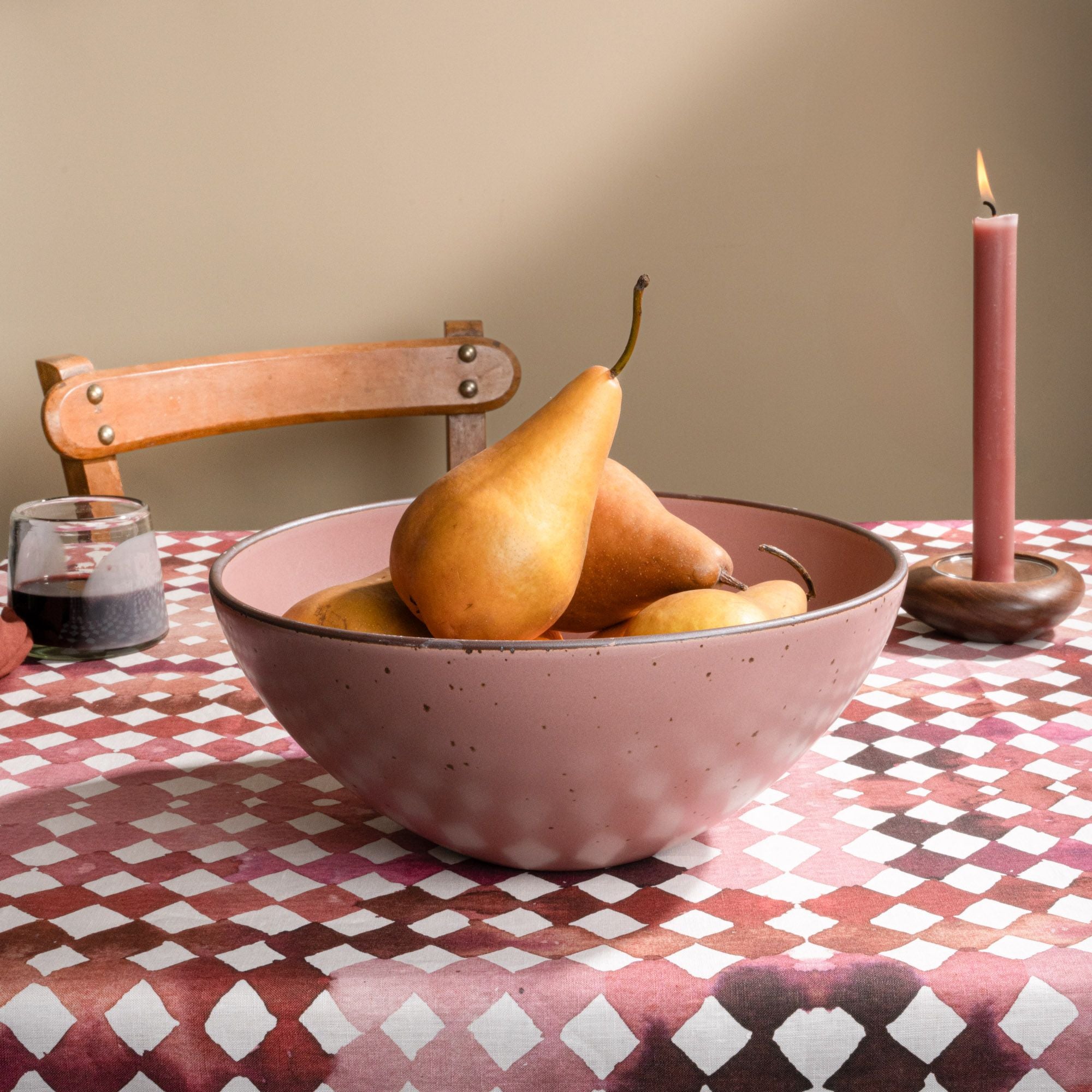 A speckled pink ceramic bowl filled with pears sits on a patterned tablecloth, accompanied by a glass of red wine and a lit pink candle in a wooden holder. A wooden chair is visible in the background.