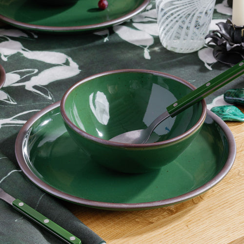 A medium rounded ceramic bowls in a glossy emerald green atop a dinner plate, with a silver and green handled spoon sitting in the bowl, part of a table setting