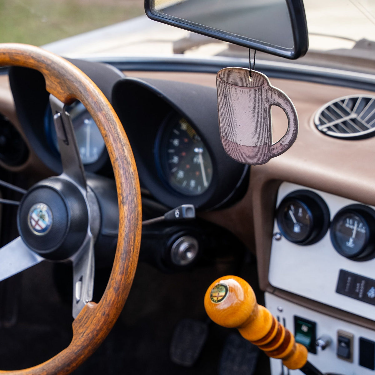 Pale brown mug-shaped East Fork air freshener hanging from a rearview mirror inside a vintage car with a wood steering wheel and classic dashboard.