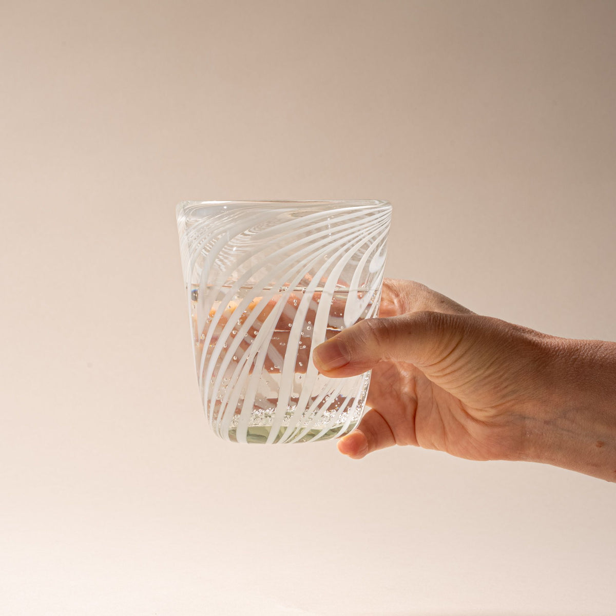 A hand holds out a clear glass tumbler with white diagonal swirl pattern against a neutral background.