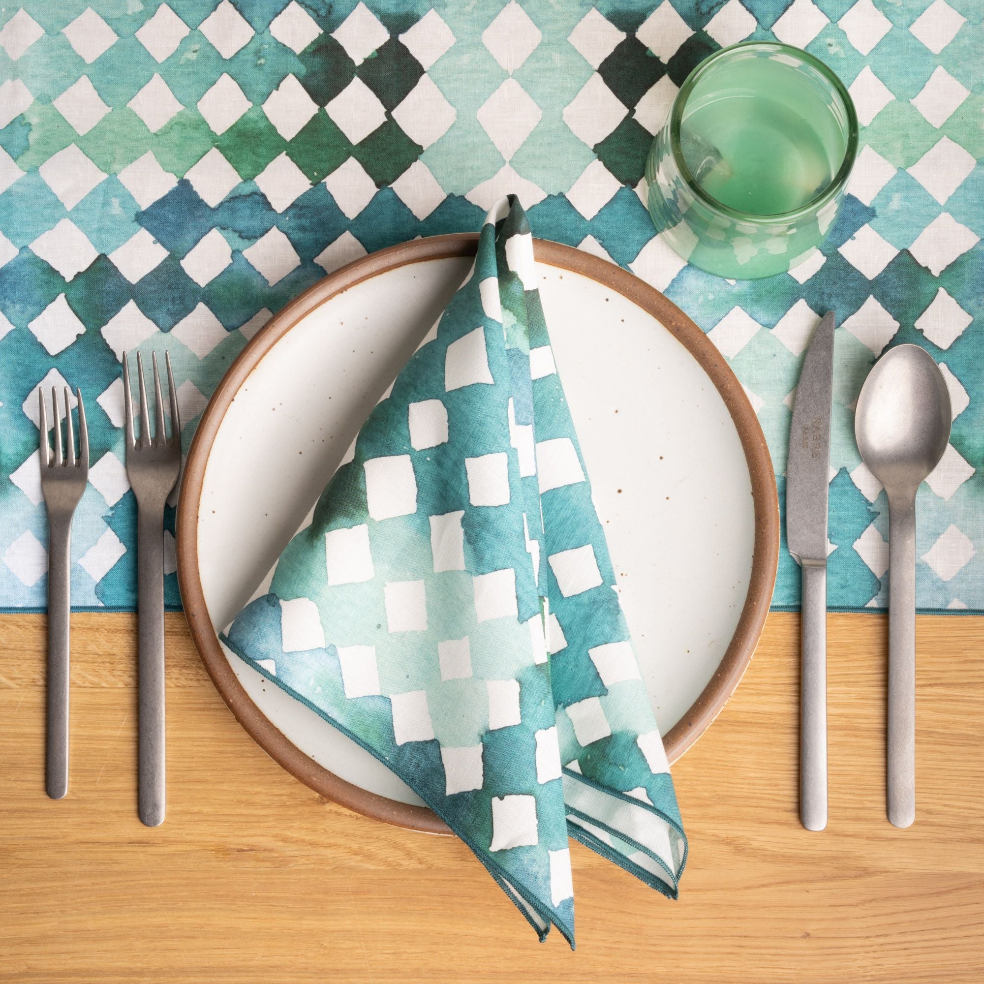 A white ceramic dinner plate with a folded watercolor diamond patterned napkin, set with silver utensils and a green glass on a matching tablecloth.