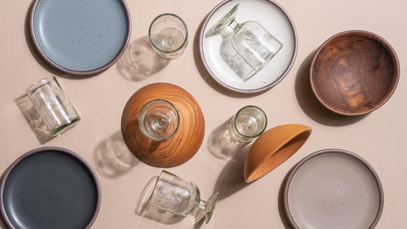 Ceramic plates, wooden bowls, and clear etched glassware arranged flat-lay on a beige surface with soft shadows.