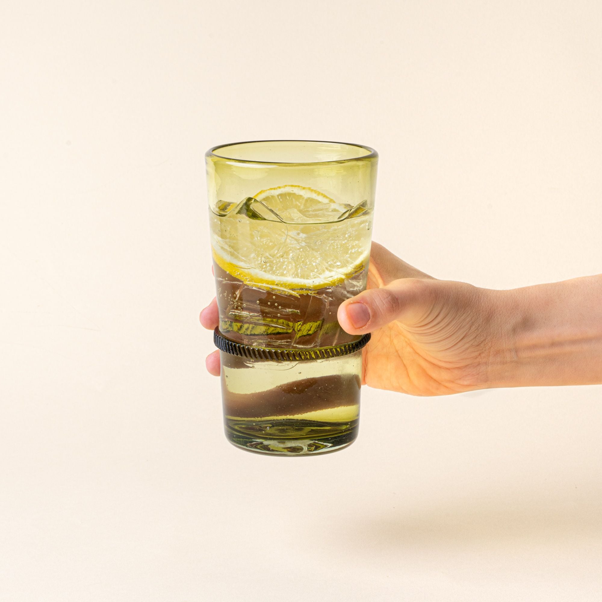 A hand holds an olive tall recycled glass tumbler with a ridged band detail, filled with water and a lemon slice.