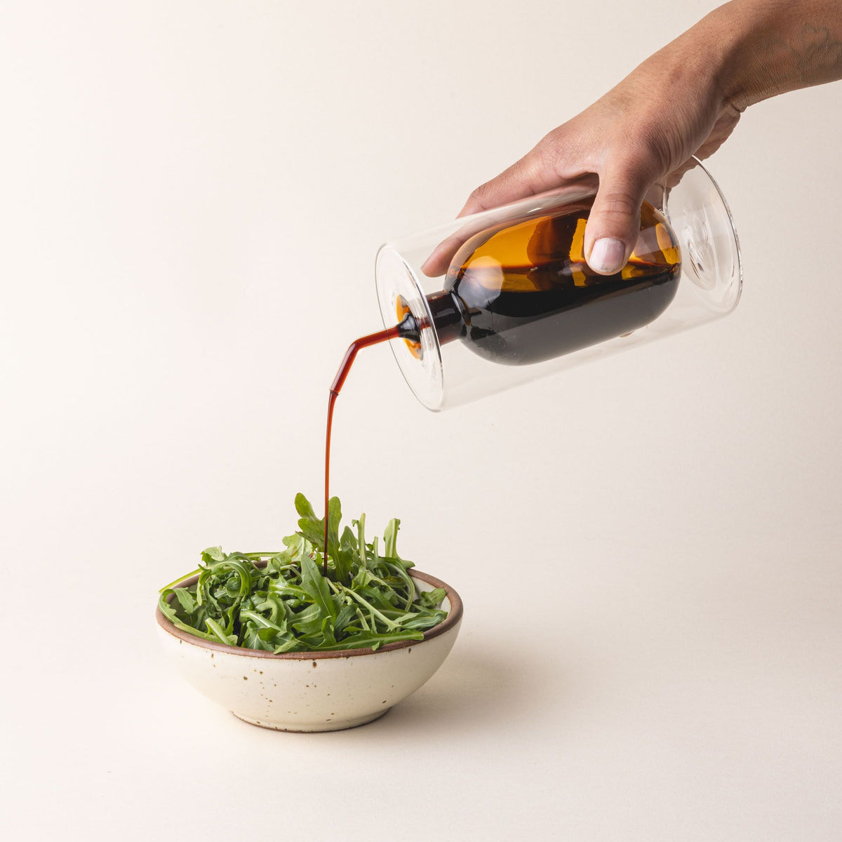 A hand holds an artful glass cruet in an orange color and is pouring it over a small side salad. There is a rounded bulb that acts as a container for the cruet, surrounded by a doubled glass wall. The cruet is filled with oil.