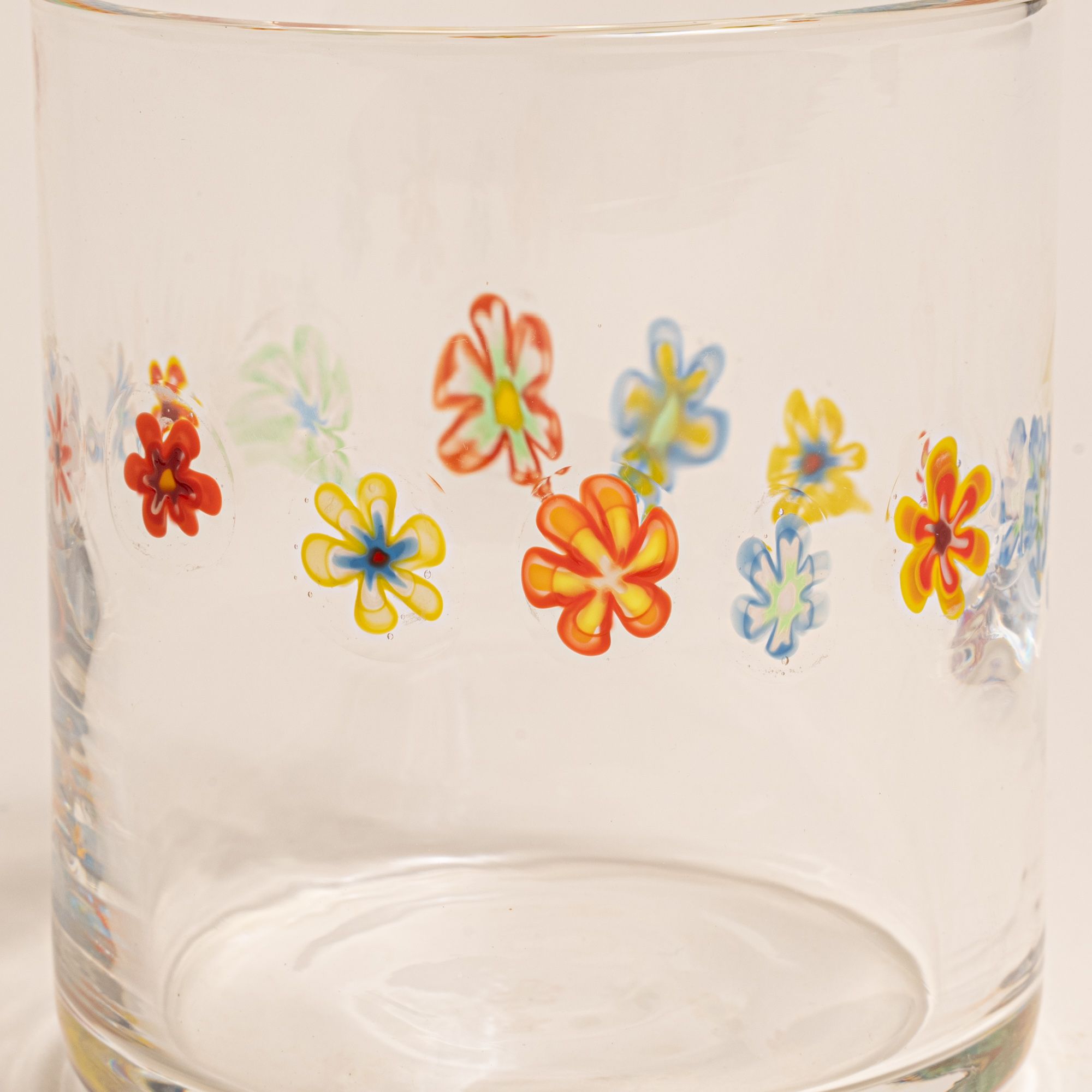 A closeup of a clear glass with a ring of multicolored little flowers on a cream background.