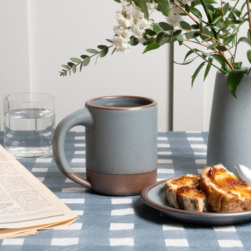 A large mug in a blue-grey color with unglazed base and rim, next to a plate with toast and newspaper.