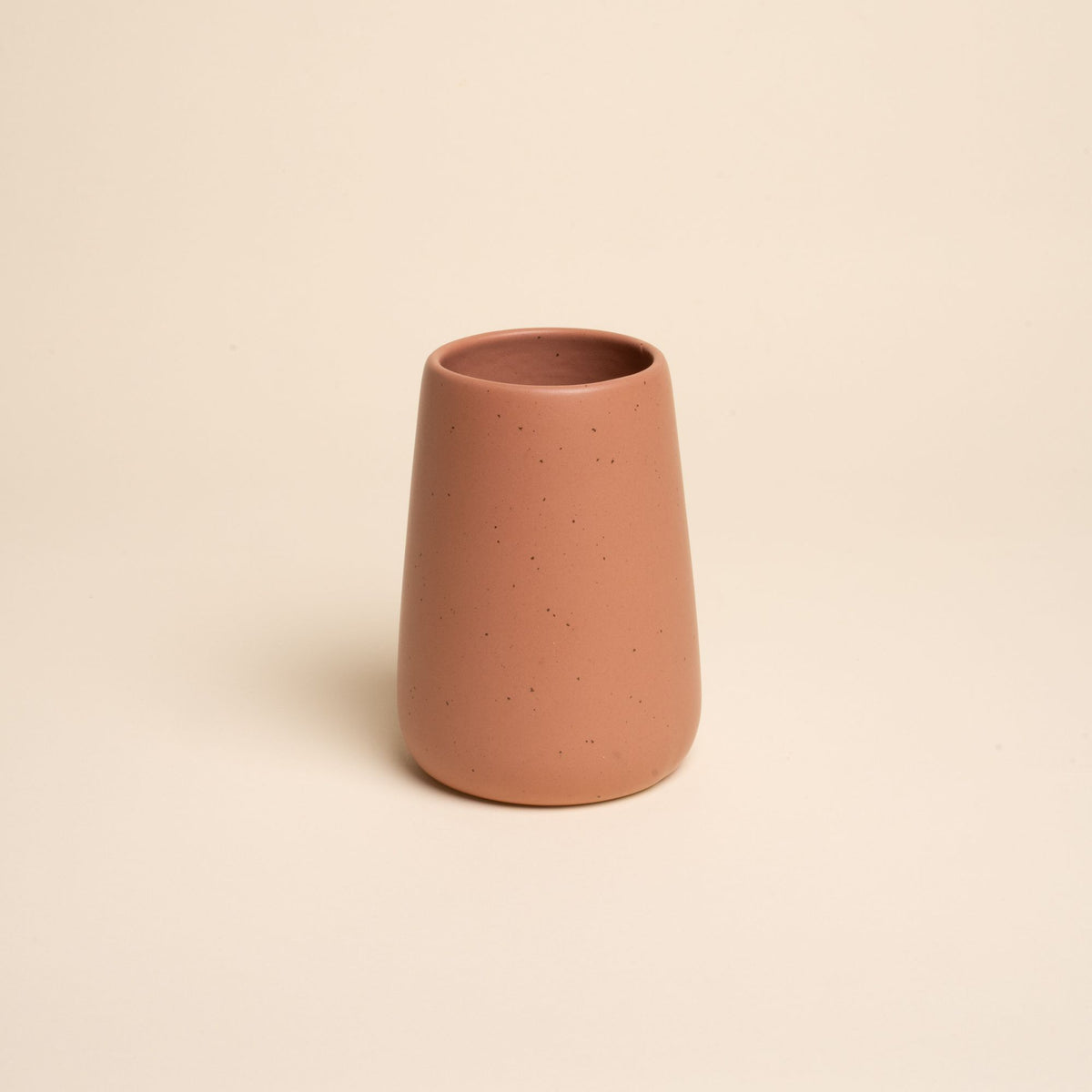 A small minimalist terracotta tumbler vase with subtle iron speckles stands centered on a soft beige background