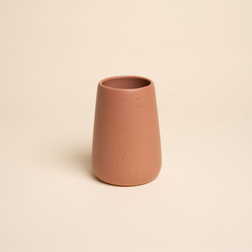A small minimalist terracotta tumbler vase with subtle iron speckles stands centered on a soft beige background
