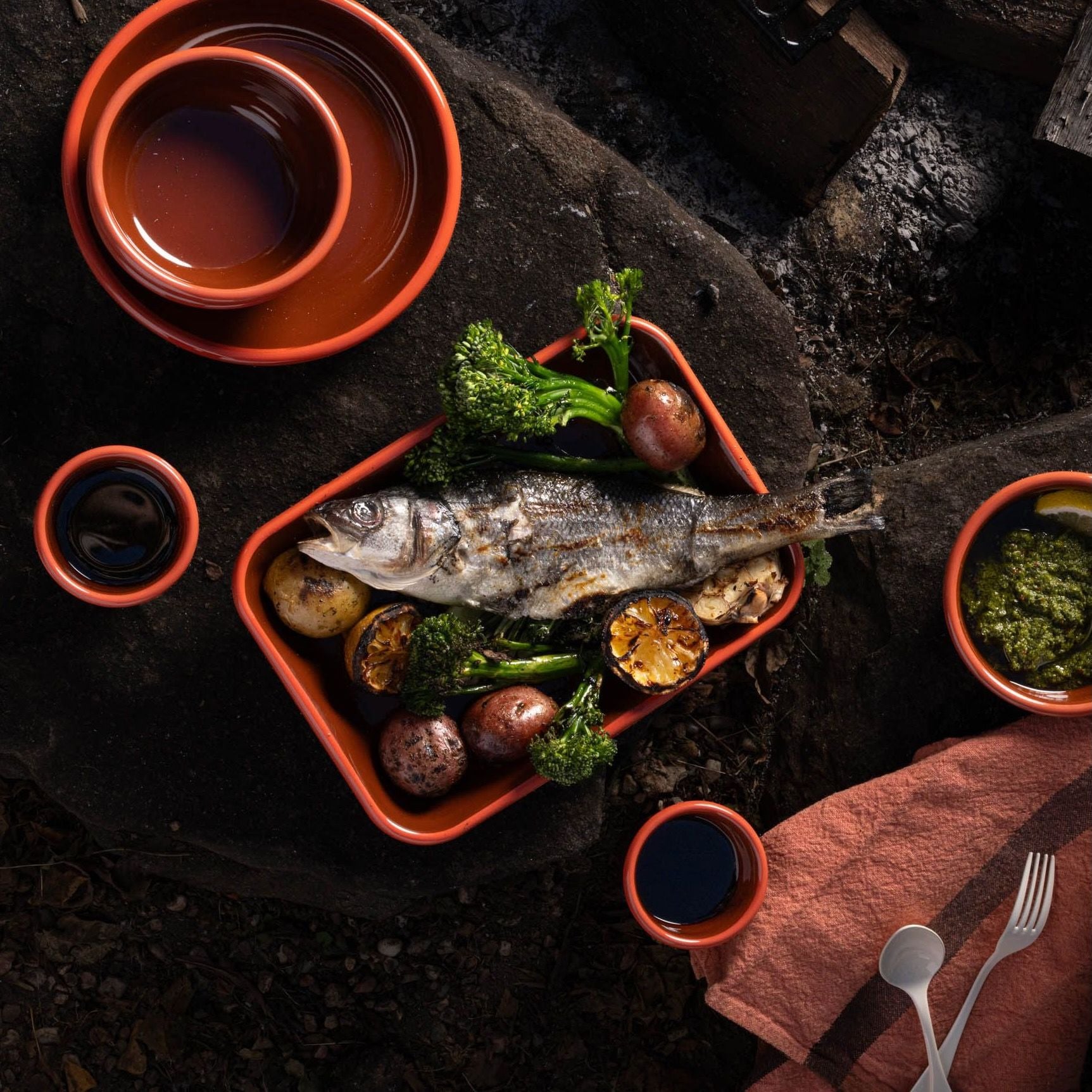 A large enamel baking dish in red-broen color filled with a roasted whole fish, potatoes and vegetables, surrounded by enamel plates and bowls of the same color.