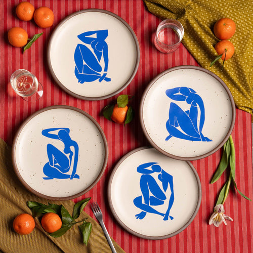 Four ceramic dinner plates in warm off-white colors and unglazed rim, each with a different blue decal from the Nu Bleu series by Henri Matisse. The plates are sitting on a red tablecloth with silverware, flowers, oranges, and glasses.