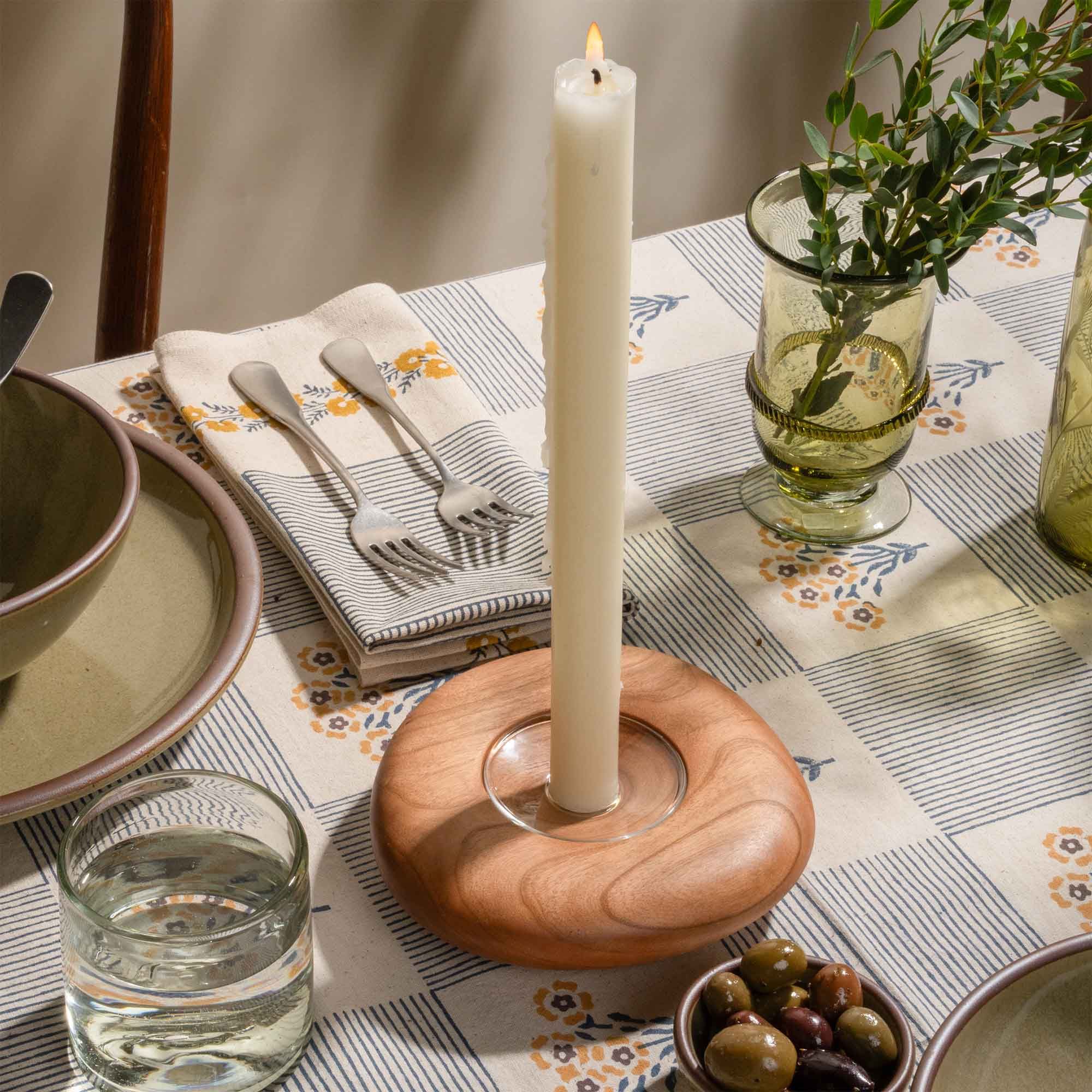 Lit white taper candle in a wooden holder on a patterned table setting with glassware and olives.