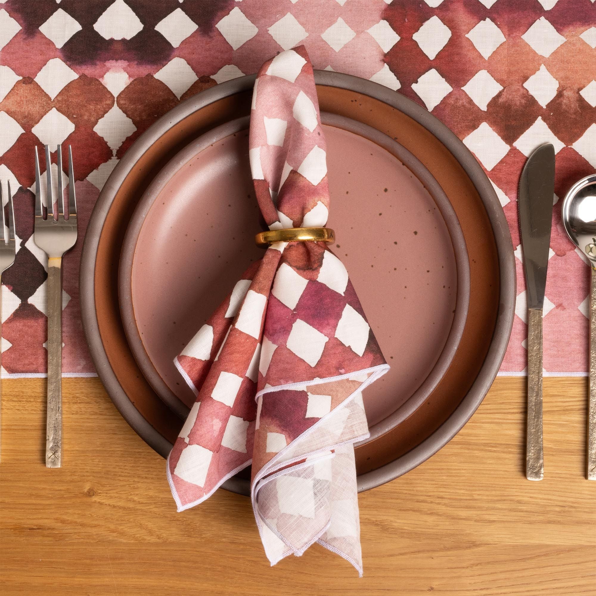 Stacked pink ceramic plates with a watercolor diamond patterned napkin and brass ring, set with silver utensils on a matching tablecloth.