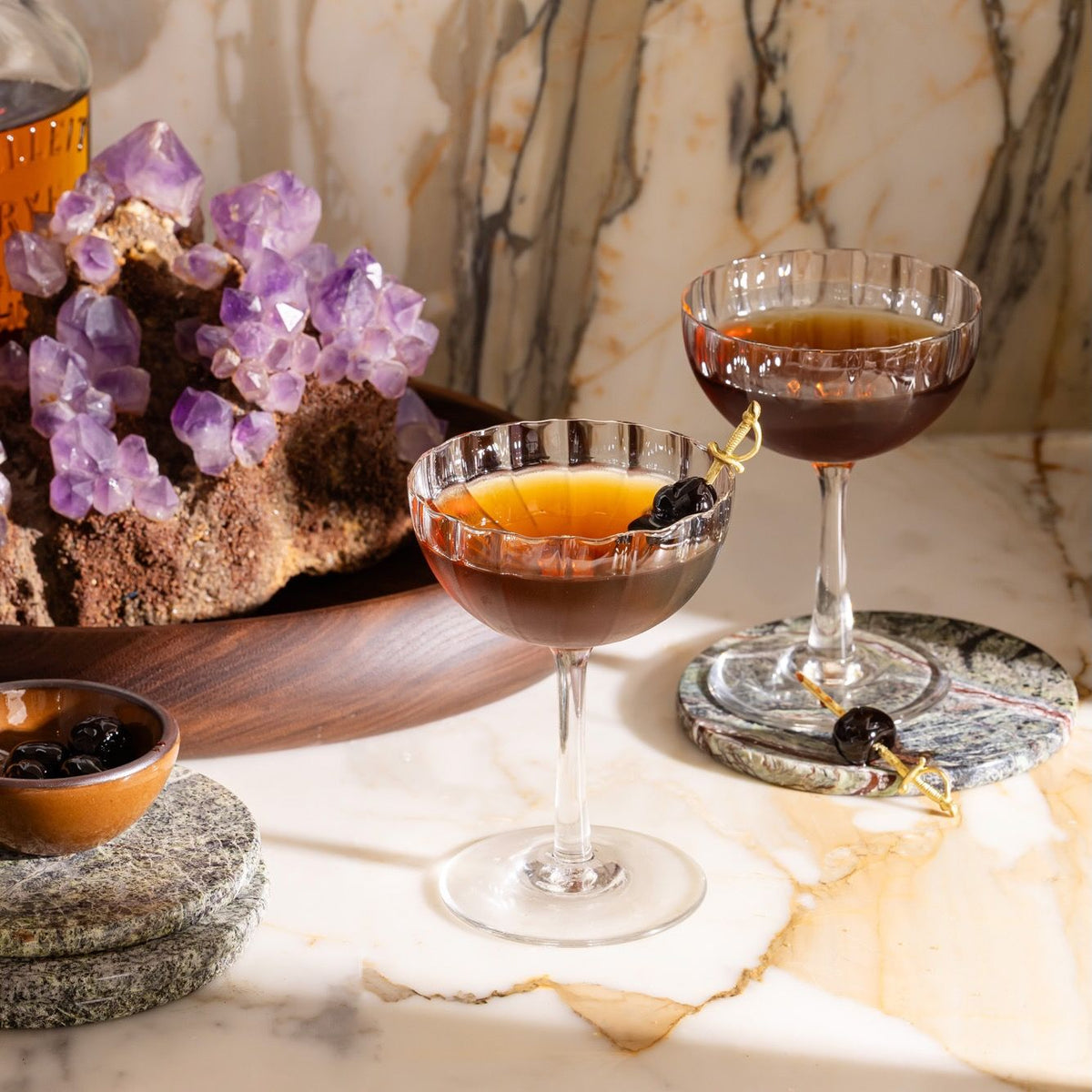 Two cocktails in coupe glasses garnished with cherries on brass sword picks, set on marble with amethyst crystals nearby.