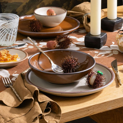 Glossy caramel brown and white bowls with rambutan, lychee, honeycomb, and candles on a patterned table setting.