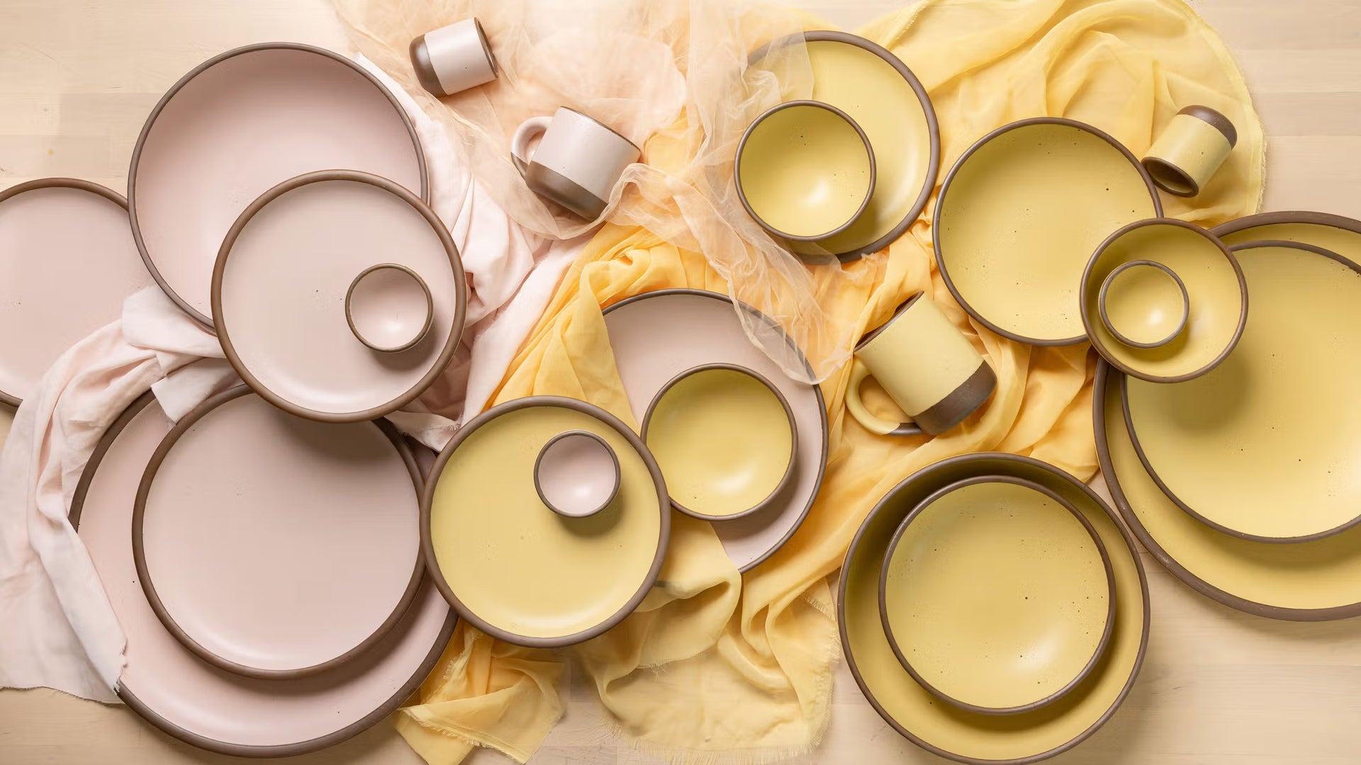 Assorted handmade ceramic plates, bowls, and mugs in soft pink and yellow glazes with brown rims, arranged on pale pink and yellow fabric on a light wood surface.
