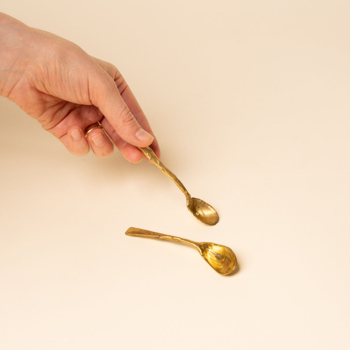 Hand holds one of two small handcrafted gold spoons with textured, organic shapes on a neutral background.