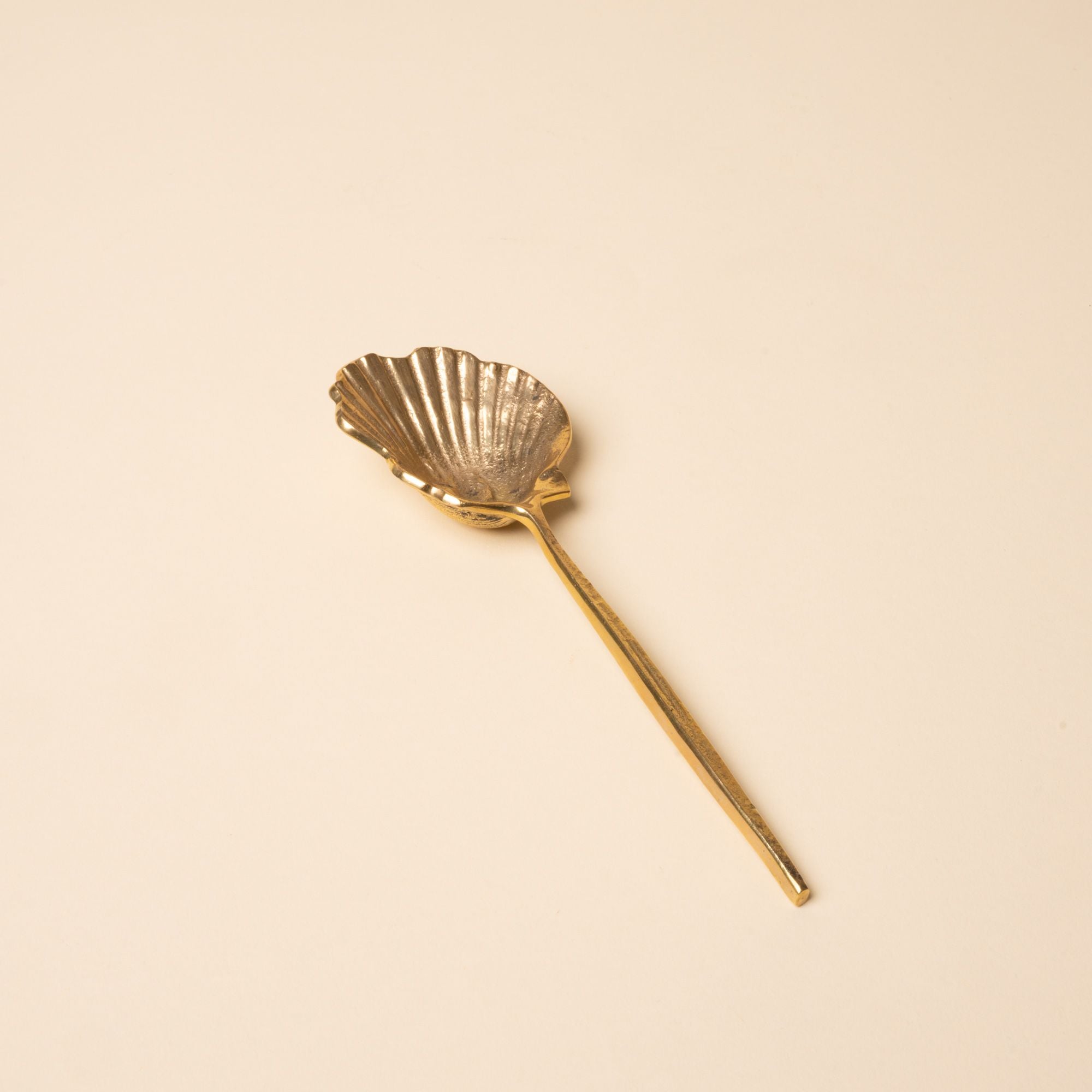 Brass spoon with a scalloped shell-shaped bowl and long handle, set against a neutral beige background.