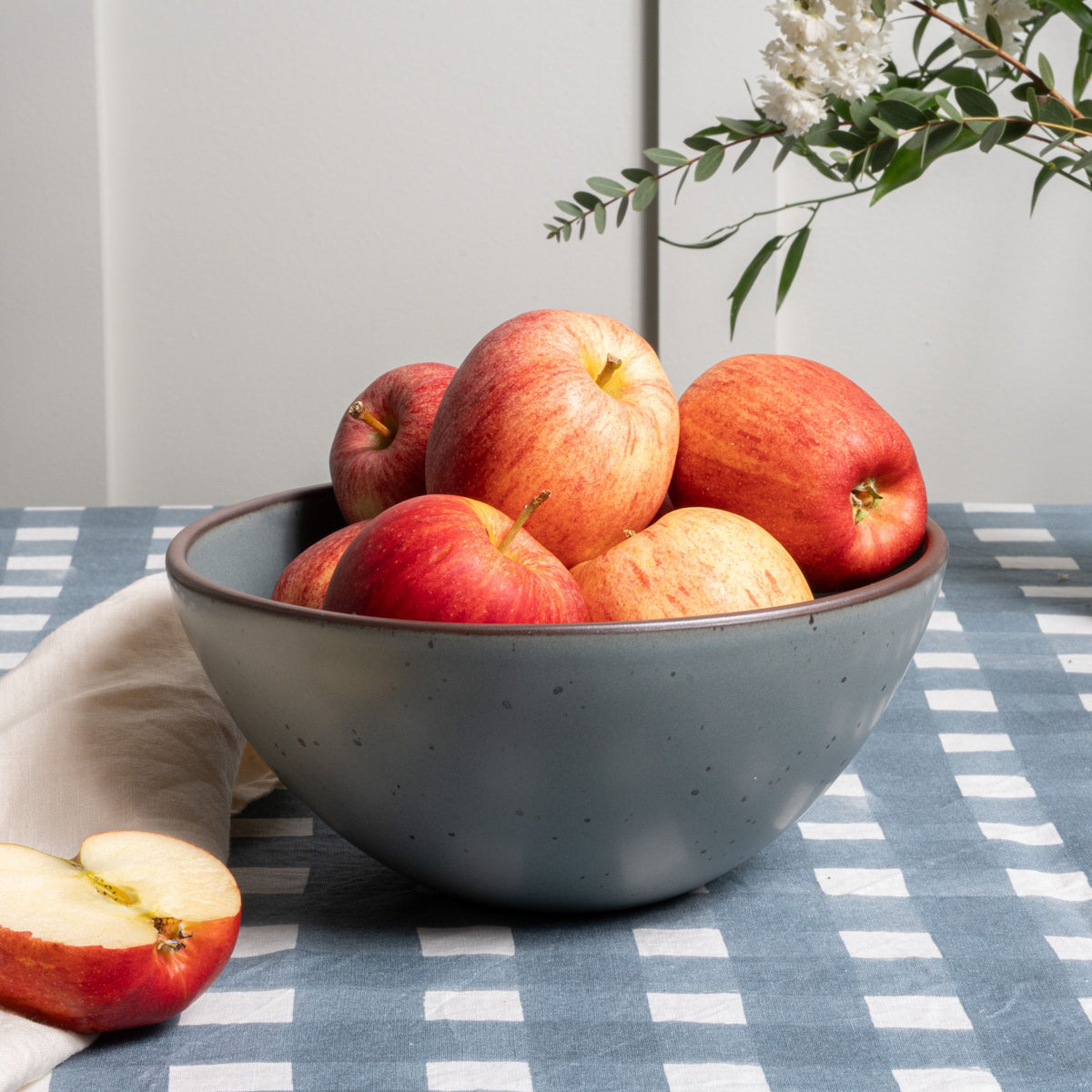 Red apples in a large ceramic mixing bowl in a blue-grey color and on a blue-grey crosshatch tablecloth