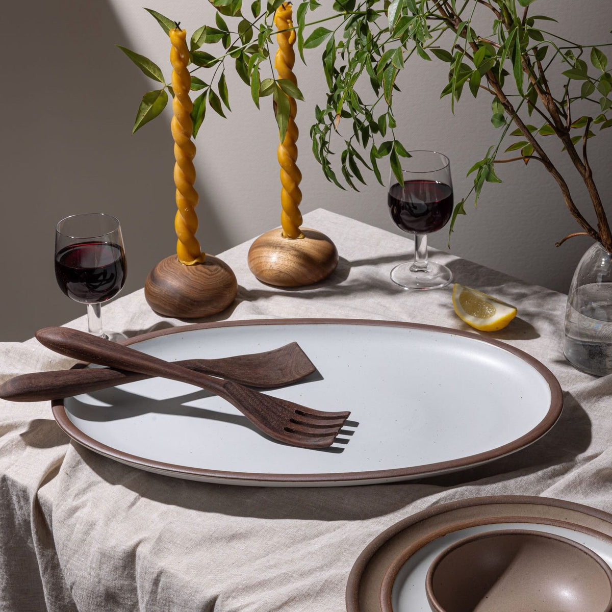 Minimal table setting with a white ceramic oval platter, wooden serving utensils, twisted beeswax candles, glasses of red wine, and greenery.
