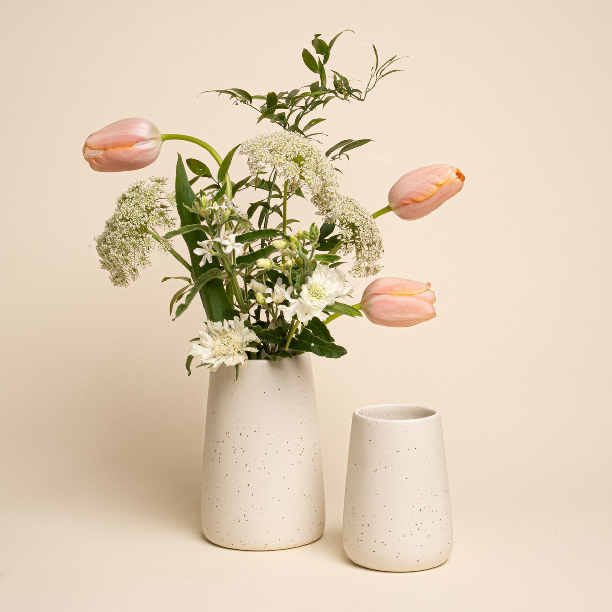 Two speckled cream vases, one filled with tulips and white flowers, styled on a neutral background.