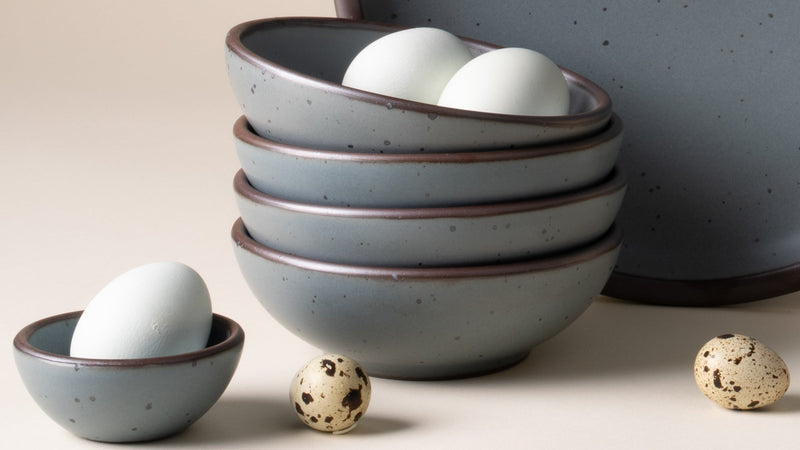Set of blue-grey ceramic bowls with speckled texture and brown rim, containing eggs and quail eggs on a neutral background.