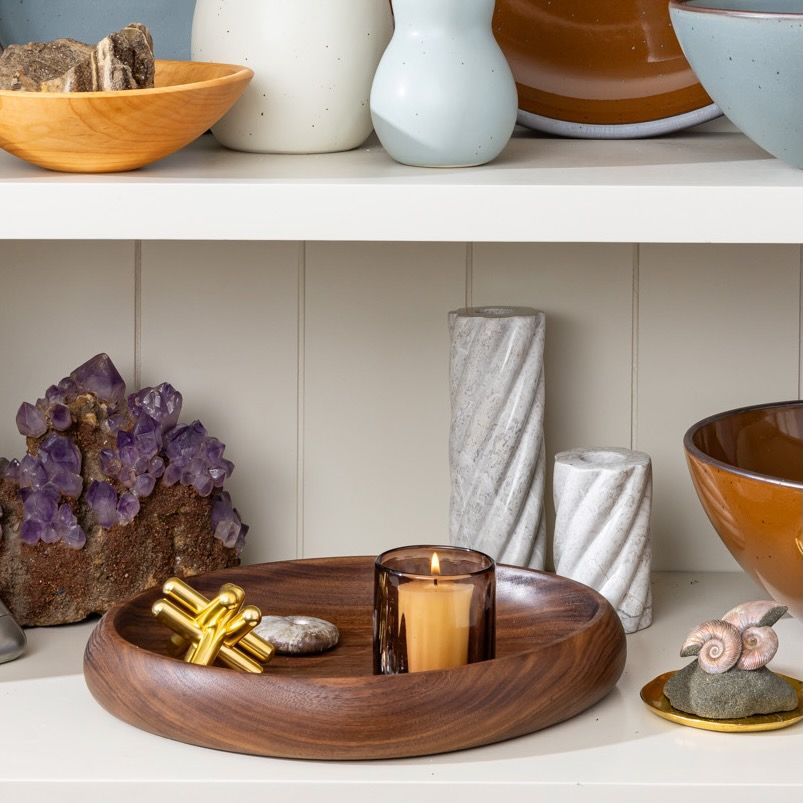 Wooden catchall tray holding a lit amber glass candle, styled on a shelf with ceramics, crystals, and stone decor.