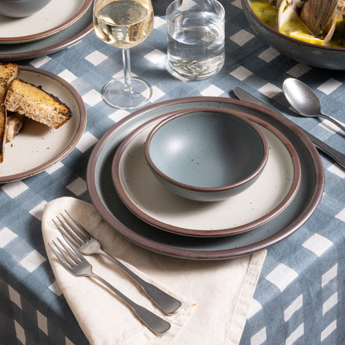 Small shallow bowl and dinner plate in a blue-grey color with a cream side plate, with silverware on a blue-grey crosshatch tablecloth