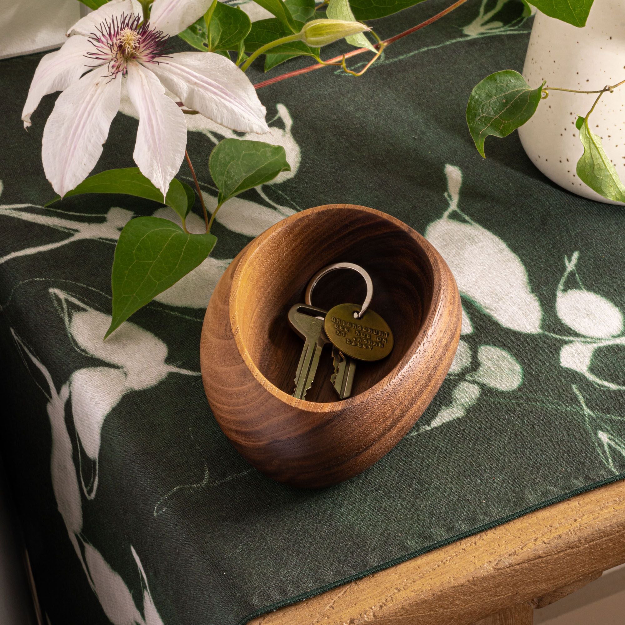 Small round walnut bowl holding house keys, placed on a green floral table runner beside a white flower and leafy plant.