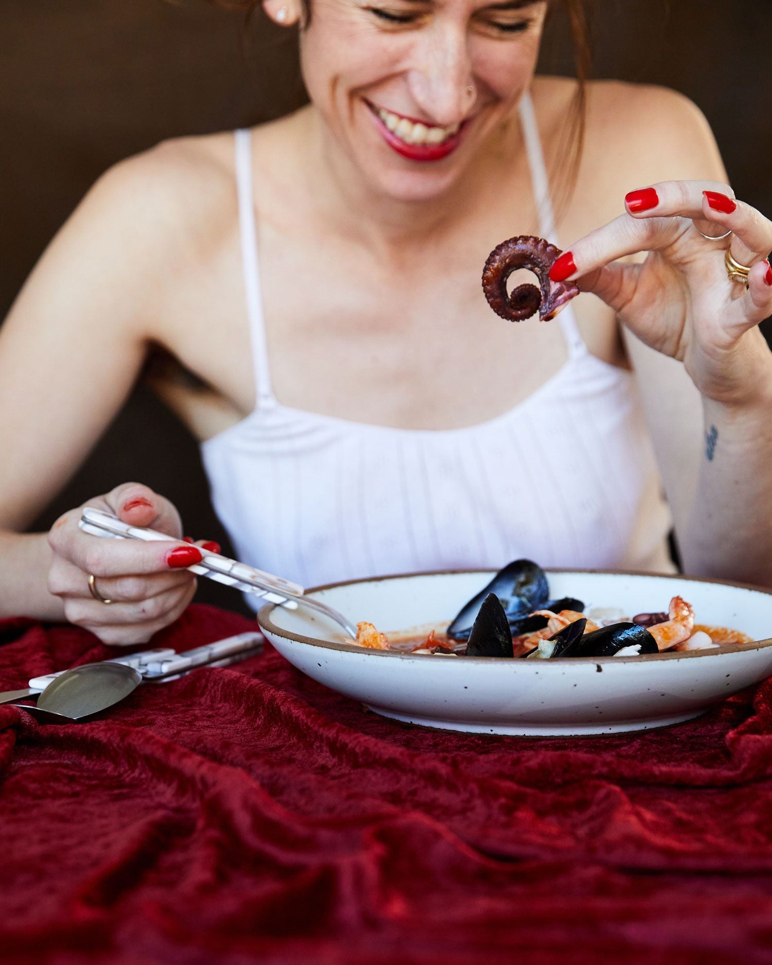 A person is smiling and has 1 hand holding a utensil on a white coupe plate, and the other holding a mussel