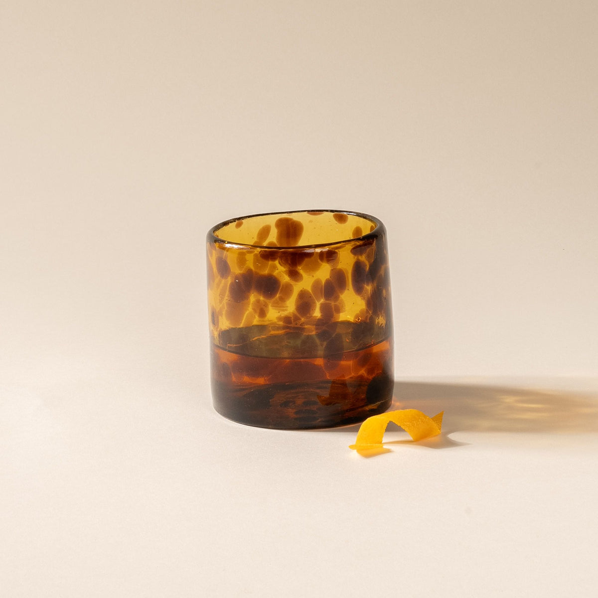 A tortoiseshell short transparent drinking glass, filled with a cocktail and an orange peel to the side
