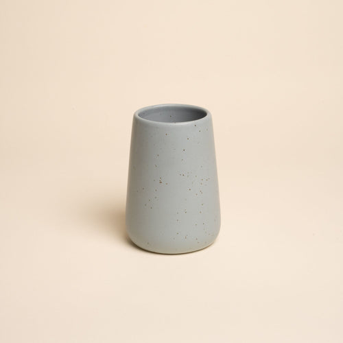 A small minimalist blue-grey tumbler vase with subtle iron speckles stands centered on a soft beige background